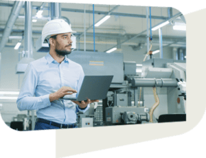 A professional man wearing a white hard hat and light blue shirt is standing in an industrial facility, holding a laptop