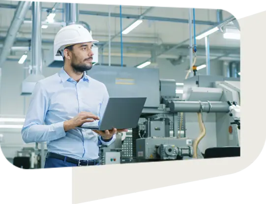 A professional man wearing a white hard hat and light blue shirt is standing in an industrial facility, holding a laptop