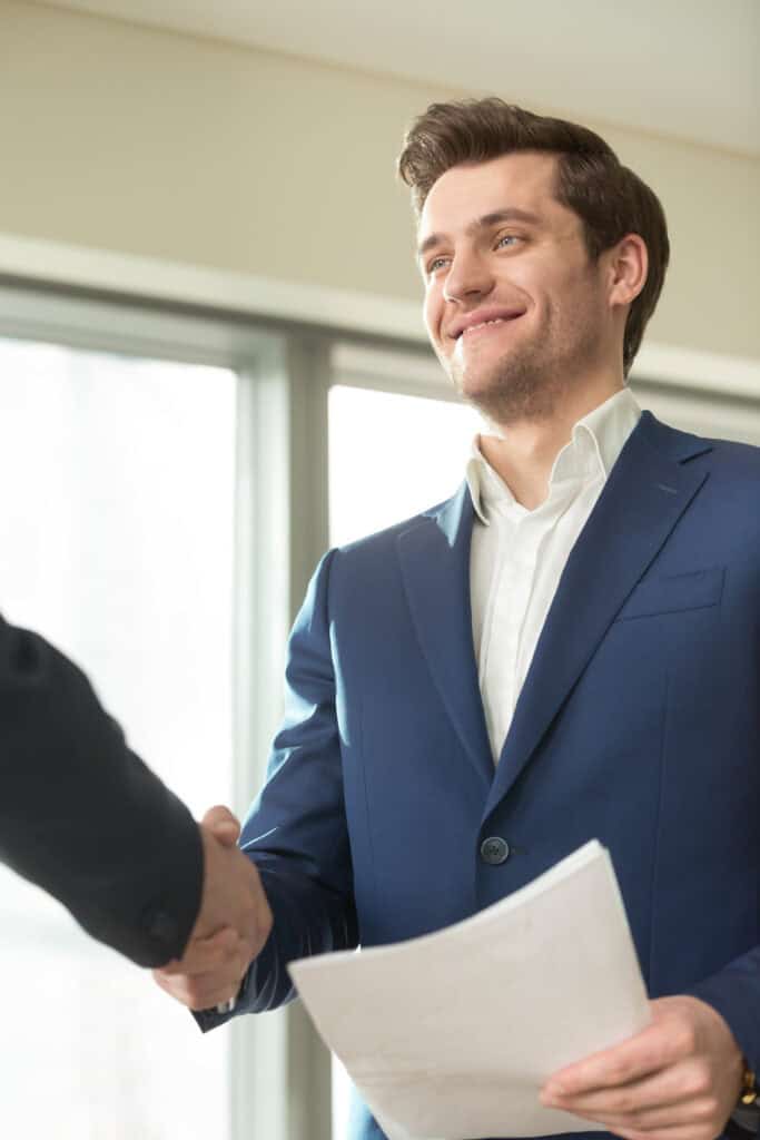 Confident smiling businessman with documents in hand handshaking with client in office