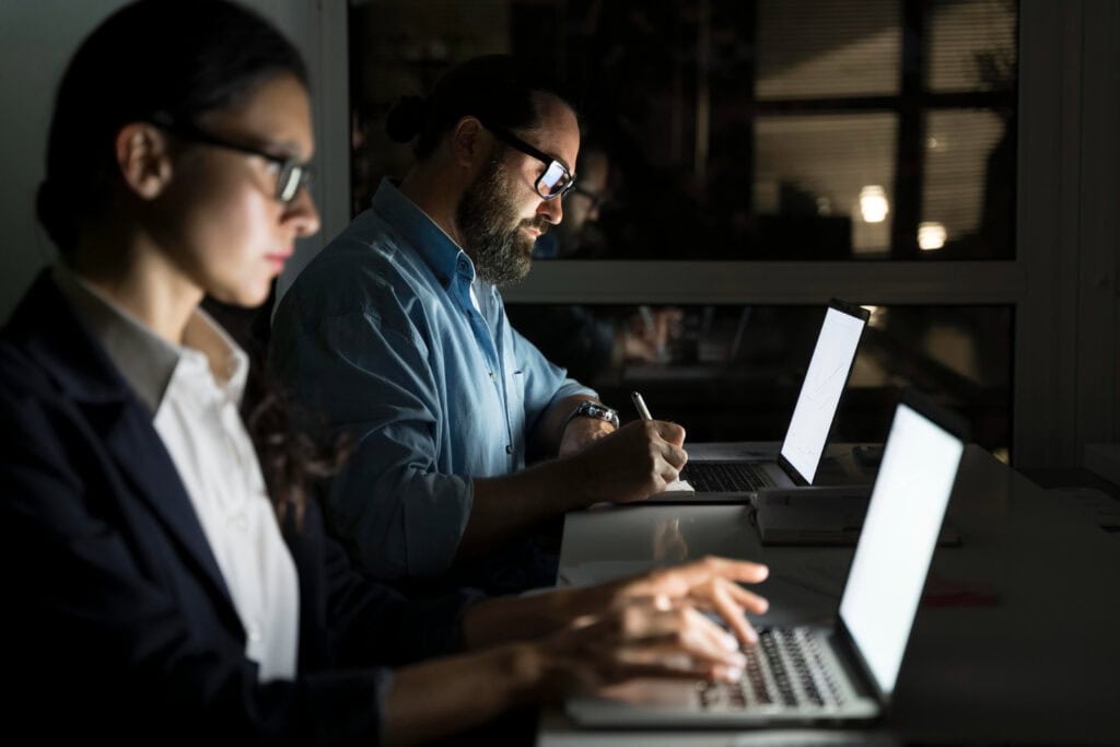 Two IT interim management experts, one woman and one man, work in dark room lit only by their laptop screens
