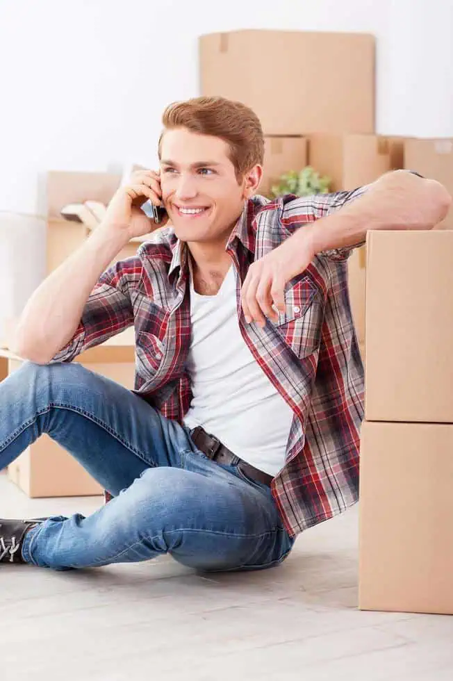 Young man sitting on the floor and talking on the mobile phone with cardboard boxes around him